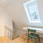 Dachgeschosszimmer mit Schreibtisch, grüner Stuhl und Dachfenster. Treppenhaus und Tür sichtbar.