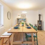 Esszimmer mit Holztisch, grünen Stühlen und Kaminofen. Fenster und Küche sind sichtbar.