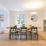 Essbereich mit Holztisch, grünen Stühlen und Fenster. Glaswand-Schrank und runder Spiegel an der Wand.