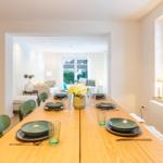 Esszimmer mit Holztisch und grünen Stühlen. Durchgang zum Wohnzimmer mit Sofa und Fenster.