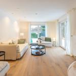 Wohnzimmer mit Sofa, Sessel und Glas-Tisch. Holzboden und große Fenster.