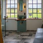 Badezimmer mit grünem Waschbecken, Holzunterschrank und großen Fenstern mit Blick auf Grünland.