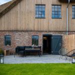 Haus mit Holzfassade und Dachstuhl, Terrasse mit Sitzgelegenheiten und Grasfläche.