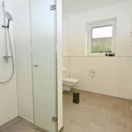 Bad mit Dusche, Toilette und Fenster. Fliesenboden und weiße Wände.