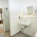 Badezimmer mit Waschbecken, Spiegel und Toilette. Durchgang zu Wohnbereich mit Holzboden.