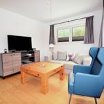 Wohnzimmer mit Sofa, blauem Sessel und Holztisch. Fernseher und Fenster mit Vorhängen.