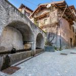 Steinerner Brunnen in einer gepflasterten Gasse mit traditionellen Häusern.