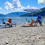 Steiniger Strand mit Liegen, Sonnenschirm und Blick auf das Wasser und Berge.