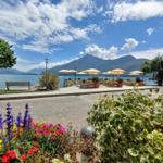 Terrasse mit Tischen, Sonnenschirmen und Blick auf den See und Berge.