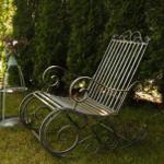 Schwungvolle Metall-Schaukelstuhl im Garten neben einer Pflanzentonne.