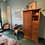 Zimmer mit Bett, Holzschrank, zwei Stühlen und Fenster.