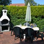 Terrasse mit Tisch, Stühlen, Sonnenschirm und Grillofen. Hintergrund: grüne Hecke.