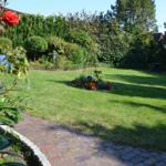 Garten mit Rasen, Blumenbeete und Steinweg. Grillbereich im Hintergrund.