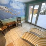 Raum mit Strandmotivwand, zwei Stühlen, Tisch und Liegestühlen. Durchgang zur Terrasse mit Blick auf Schnee.