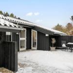 Black wooden house with snow-covered roof and garden area with table and chairs.