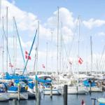 Hafen mit Segelbooten und dänischen Flaggen unter blauem Himmel.