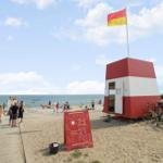 Strand mit Rettungshütte, Fahne und Fahrrädern. Besucher genießen das Wasser.