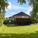 A house with a balcony stands on a green lawn surrounded by trees.