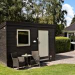 Wooden house with terrace, table, and chairs. Surrounded by greenery and sky.