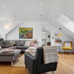Wohnzimmer mit grauem Sofa, schwarzer Ledercouch und Holzboden unter schrägen weißen Wänden.