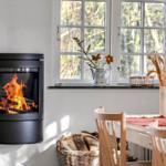 Wohnraum mit Kaminofen, Tisch und Stühlen, Fenster mit Blumen und Licht.