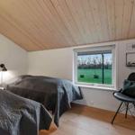 Zwei Betten mit grauen Bettdecken, Holzdecke, Fenster mit Blick auf Grünfläche und schwarze Sitzgelegenheit.