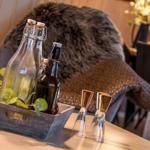 Holztablett mit Flaschen und Gläsern auf Tisch, Hintergrund mit Felldecke und Blumen.
