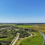 Weitläufige Landschaft mit Wiesen, Wald und einem Fluss. Windräder sind im Hintergrund sichtbar.