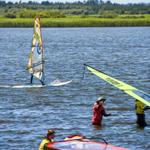 Windsurfer üben auf ruhigem Wasser. Grün bewachsener Uferstreifen im Hintergrund.