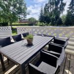 Terrasse mit Tisch und sechs Stühlen, weißer Zaun, Blick auf Grünflächen und Bäume.