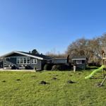 Ein graues Ferienhaus mit großem Garten und Kinderspielplatz unter blauem Himmel.
