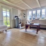 Wohnzimmer mit Sofa, Kaminofen und Holzboden. Große Fenster und Türen mit Blick auf Garten.