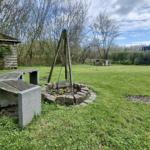 Grüner Garten mit Steinbank, Brunnen und Holzschuppen. Im Hintergrund steht ein Gebäude.
