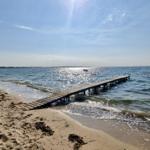 Holzsteg führt vom Strand ins Meer. Boot liegt im Wasser. Sonne scheint.