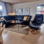 Wohnzimmer mit schwarzer Ledercouch, Sessel und Holzboden. Fenster und Tür bieten Blick nach draußen.