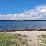 Strand mit Sand und Gras, See mit Segelboot und Boot im Hintergrund unter blauem Himmel mit Wolken.