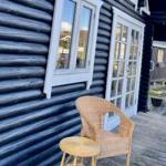 Schwarze Holzhausfassade mit weißen Fenstern und einem Rattanstuhl auf der Terrasse.