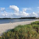 Strand mit Sand, Gras und Wasser unter blauem Himmel mit Wolken.