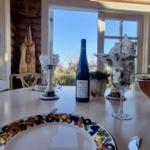 Tisch mit Teller, Weinflasche und Gläsern vor Fenster mit Blick auf Außenbereich.