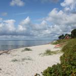 Strand mit weißem Sand, rotem Boot und kleinem Haus am Wasser.