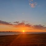 Sonnenuntergang über See mit Feld im Vordergrund und Mond am Himmel.