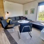 Wohnzimmer mit Sofa, Sitzgruppe und Kamin. Große Fenster mit Blick auf Grünfläche.