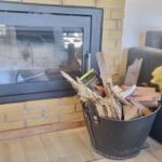 Kamin mit Holz im Eimer neben einem grauen Sofa mit gelber Decke.