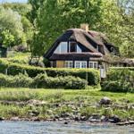 Hütte mit Strohdach am Wasser, umgeben von Grün und Bäumen.