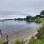 Holzsteg führt zum Wasser, Ufer mit Grünflächen und Bootsanlegestellen.