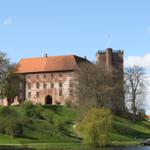 Schloss auf grüner Anhöhe mit Flagge, umgeben von Bäumen und Wasser.
