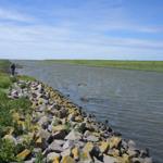 Wasserkanal mit Steinufer, Grasland und zwei Personen in der Ferne.
