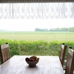 Esstisch mit Stühlen vor großem Fenster mit Blick auf Feld und Hecke.