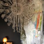 Ein Raum mit einer Glas-Kronleuchter, einer abstrakten Wandmalerei und einer kleinen Lampe auf einem Holztisch.