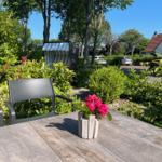 Holztisch mit Stuhl und Blumen im Garten. Im Hintergrund Bäume und ein Pavillon.